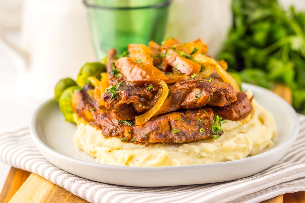 A plate of mashed potatoes topped with braised beef strips, sautéed onions, and garnished with parsley, accompanied by Brussels sprouts, served on a striped napkin.
