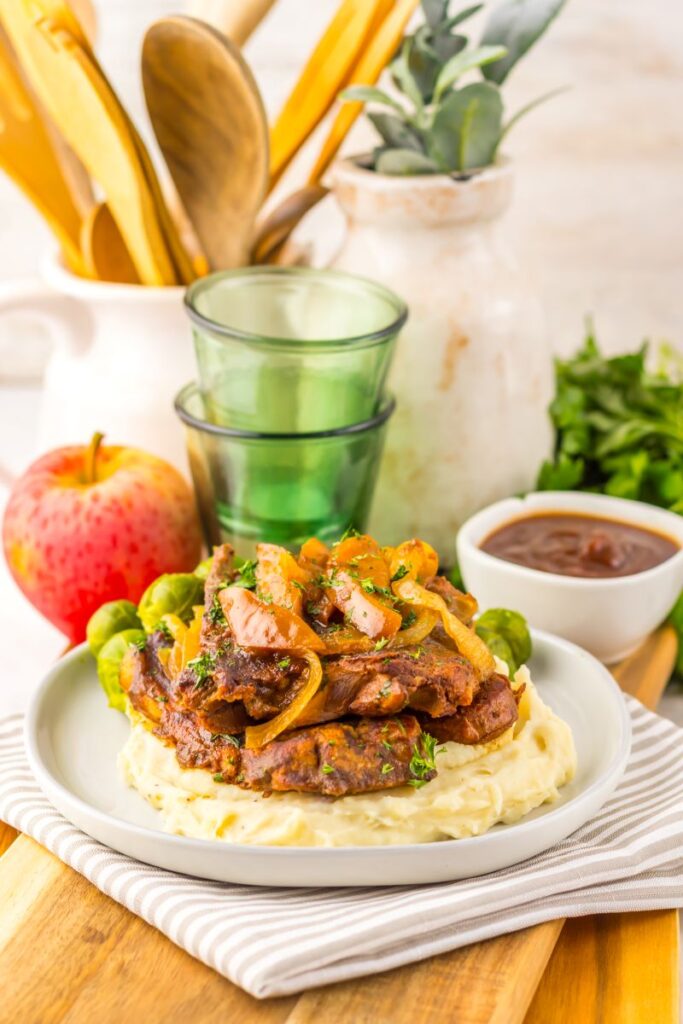 A plate of mashed potatoes topped with grilled meat, sautéed onions, and herbs, served with Brussels sprouts. In the background are an apple, green glasses, a bowl of sauce, and kitchen utensils.