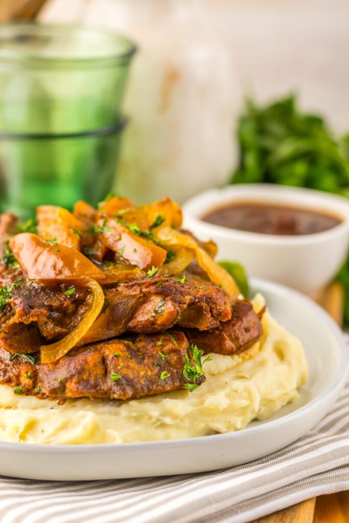 A plate of mashed potatoes topped with braised beef, sautéed onions, and fresh herbs, served with a side of brown gravy. Green glassware and herbs are blurred in the background.