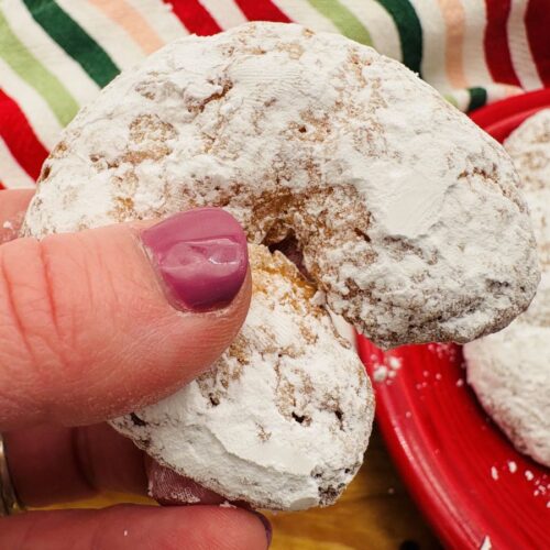 A hand with purple nail polish holds a crescent-shaped cookie covered in powdered sugar, with a red plate of similar cookies and a striped cloth in the background.