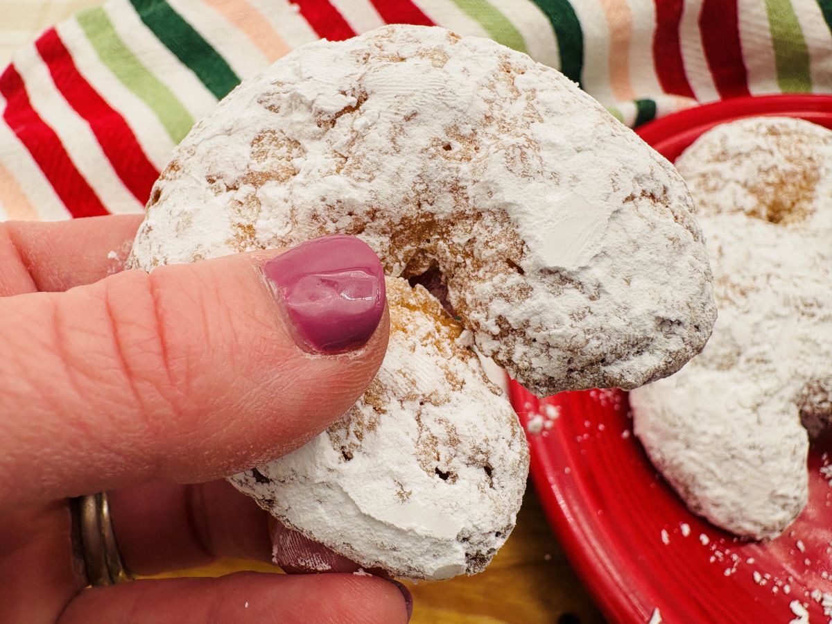 A hand with purple nail polish holds a crescent-shaped cookie covered in powdered sugar, with a red plate of similar cookies and a striped cloth in the background.
