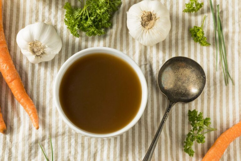 A bowl of brown broth sits on a striped cloth, surrounded by carrots, garlic bulbs, parsley, chives, and a metal ladle.