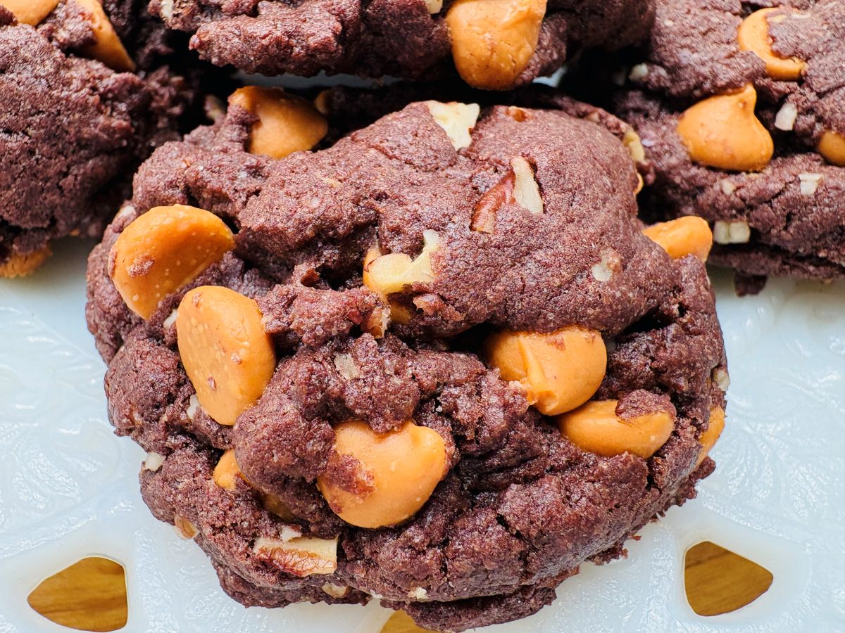 A close-up of a thick, chocolate cookie studded with large golden butterscotch chips and small pieces of nuts, resting on a white patterned surface.