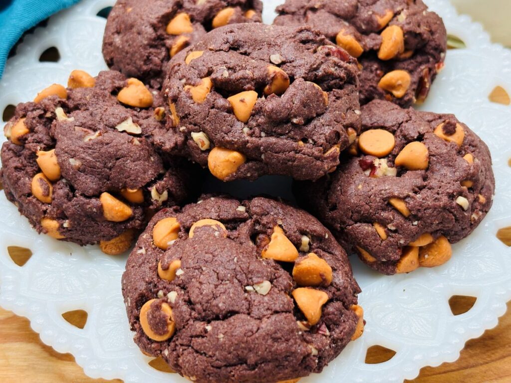 A plate of thick, chocolate cookies with golden butterscotch chips scattered throughout, arranged on a decorative white serving dish.