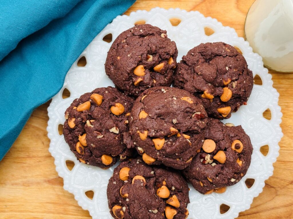 A white plate holds six chocolate cookies with peanut butter chips, on a wooden surface. A blue cloth sits to the left, and a glass of milk is partially visible in the top right corner.