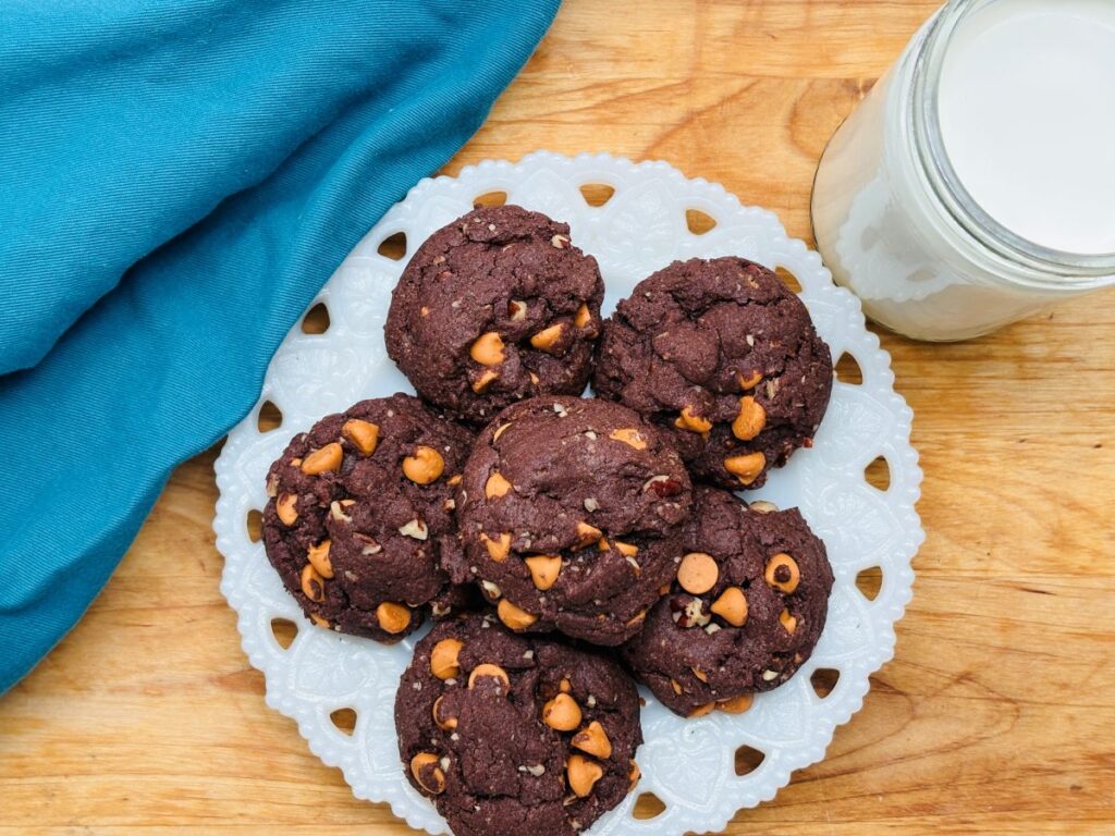 A white lace-patterned plate with six chocolate cookies studded with peanut butter chips sits on a wooden surface, next to a blue cloth and a glass jar filled with milk.
