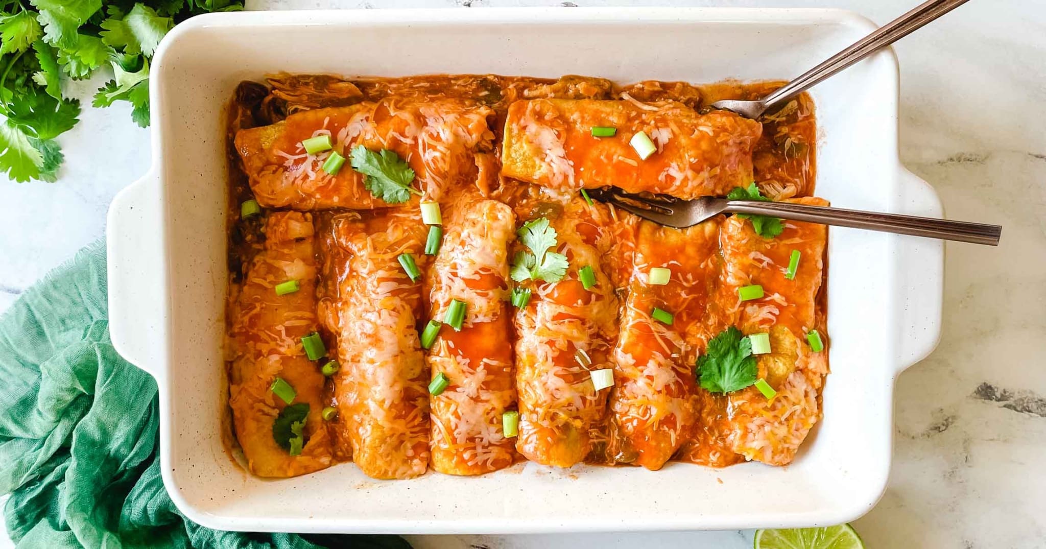 A white baking dish filled with cheesy enchiladas topped with red sauce, chopped green onions, and cilantro. Two forks are placed in the dish, and fresh cilantro and a lime slice are nearby.