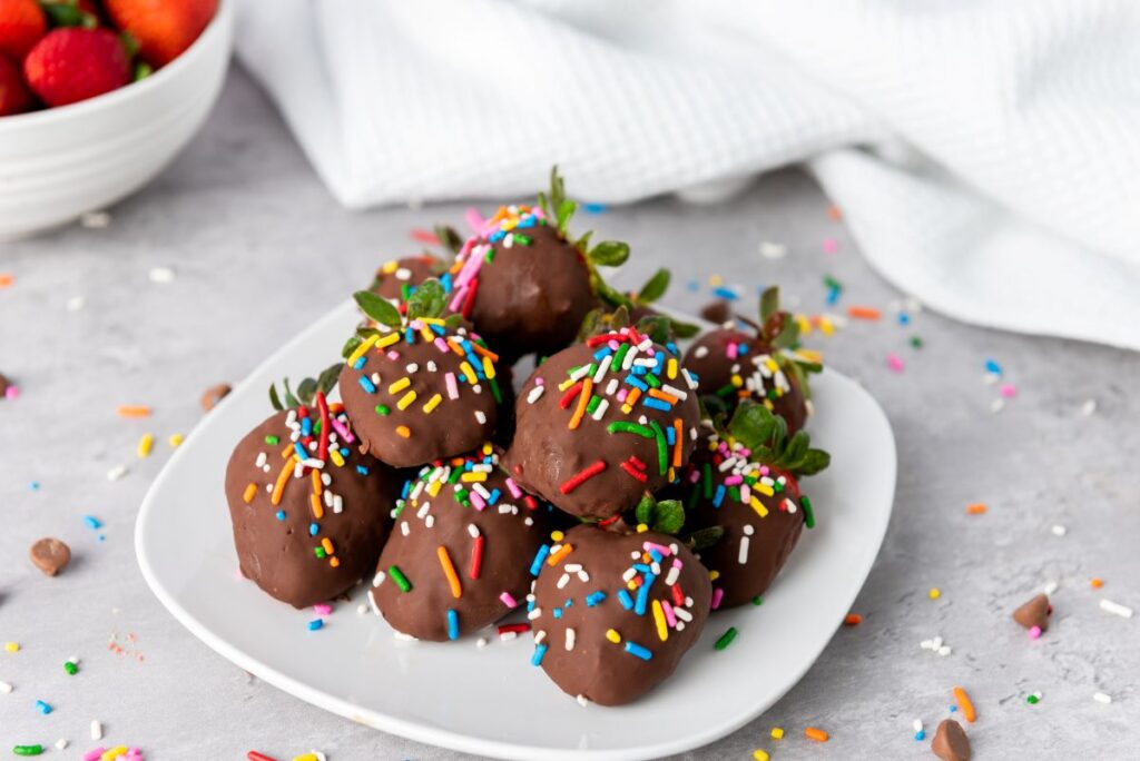 A plate of chocolate-covered strawberries topped with colorful sprinkles sits on a light surface, with a white bowl of fresh strawberries and a white cloth in the background. Sprinkles are scattered around the plate.