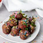 A plate of chocolate-covered strawberries topped with colorful sprinkles sits on a light surface, with a white bowl of fresh strawberries and a white cloth in the background. Sprinkles are scattered around the plate.