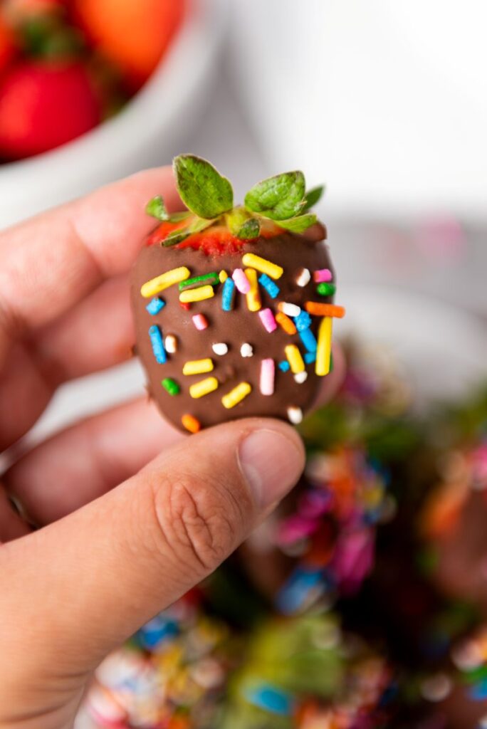 A hand holds a chocolate-covered strawberry topped with colorful rainbow sprinkles. In the background, more decorated strawberries and a bowl of plain strawberries are visible.