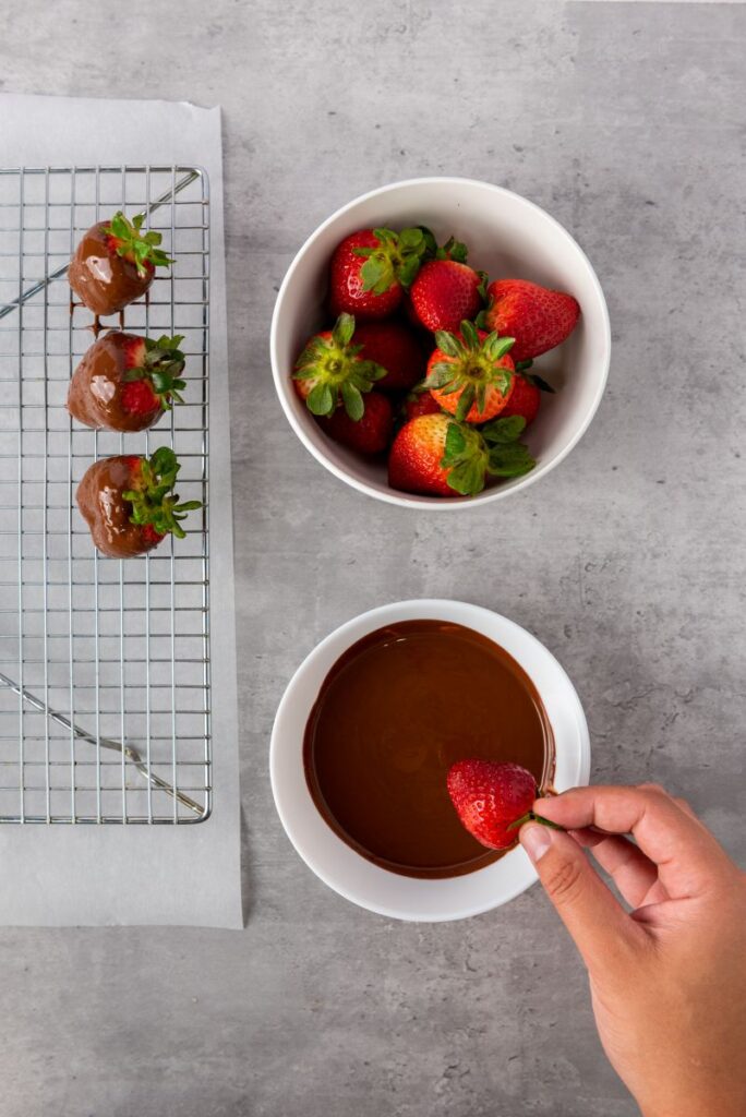 A hand dips a fresh strawberry into a bowl of melted chocolate. Next to it, a bowl is filled with fresh strawberries, and a cooling rack holds three chocolate-covered strawberries on parchment paper.