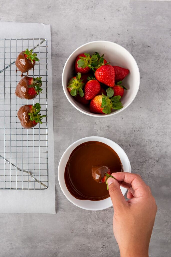 A hand dips a fresh strawberry into a bowl of melted chocolate. Nearby, a bowl of whole strawberries and a cooling rack with chocolate-covered strawberries are on a gray surface.