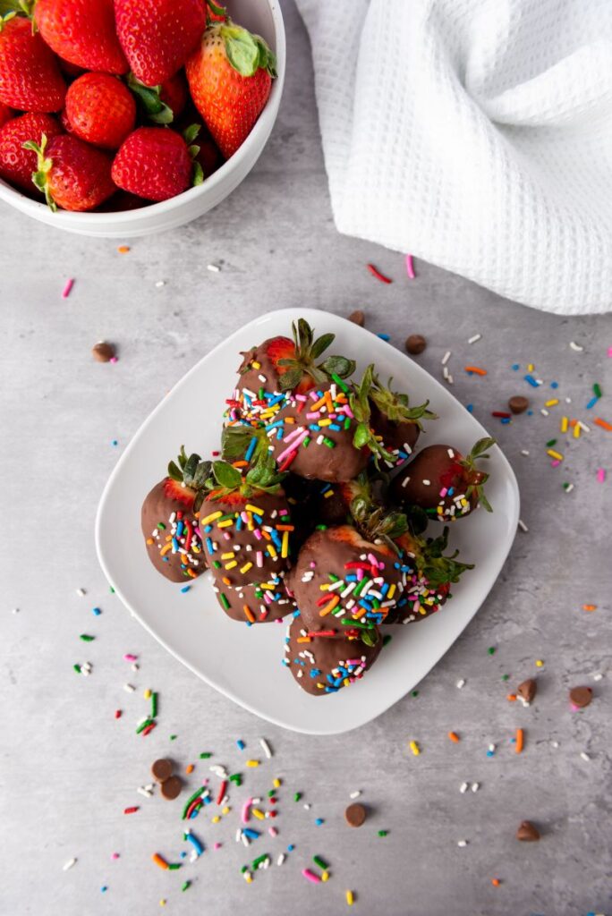 A plate of chocolate-covered strawberries topped with colorful sprinkles sits on a gray surface, with a bowl of fresh strawberries and a white cloth nearby. Sprinkles and chocolate chips are scattered around.