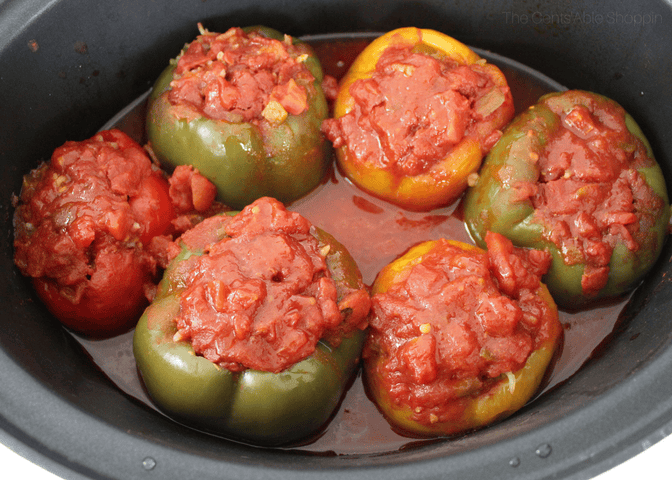 Six bell peppers—green, red, and yellow—stuffed with a tomato-based filling are arranged in a black slow cooker, surrounded by tomato sauce.