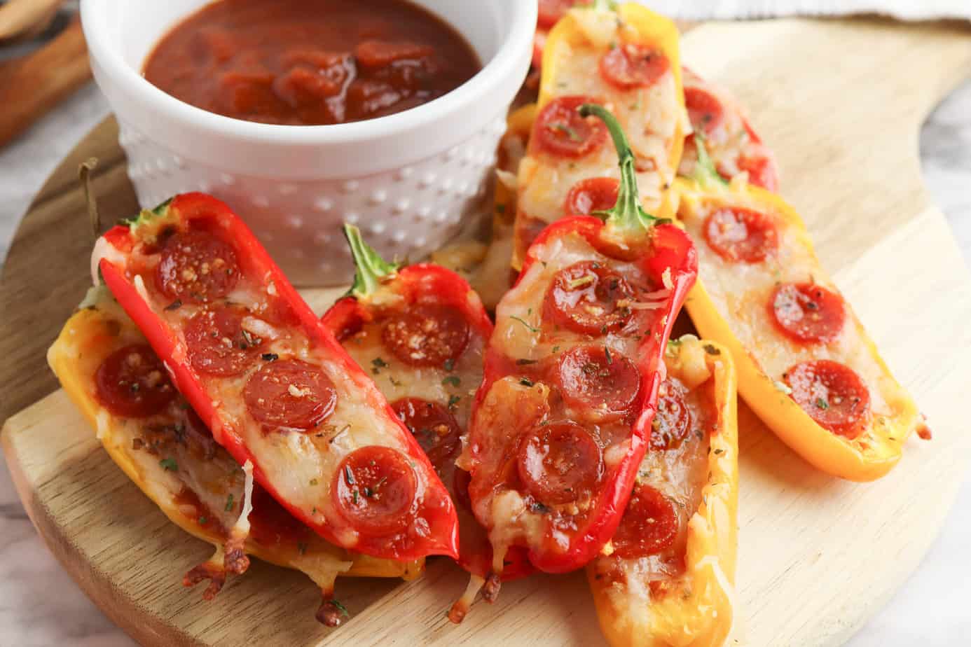 Mini bell peppers sliced in half and topped with melted cheese and small pepperoni slices, arranged on a wooden board with a bowl of marinara sauce for dipping.