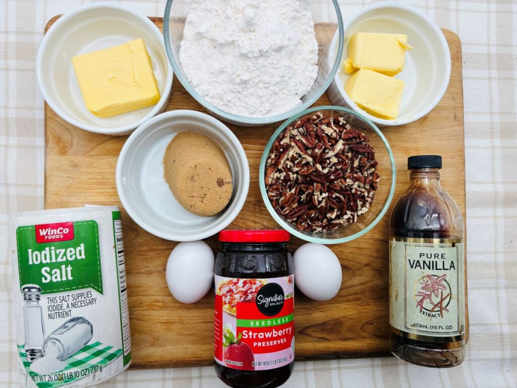 A wooden board with ingredients for baking: two bowls of butter, a bowl of flour, chopped pecans, brown sugar, two eggs, a jar of strawberry preserves, a bottle of vanilla, and a container of iodized salt.