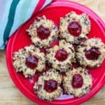 A red plate holds seven thumbprint cookies coated in chopped nuts and filled with red jam, placed on a wooden surface next to a striped towel.