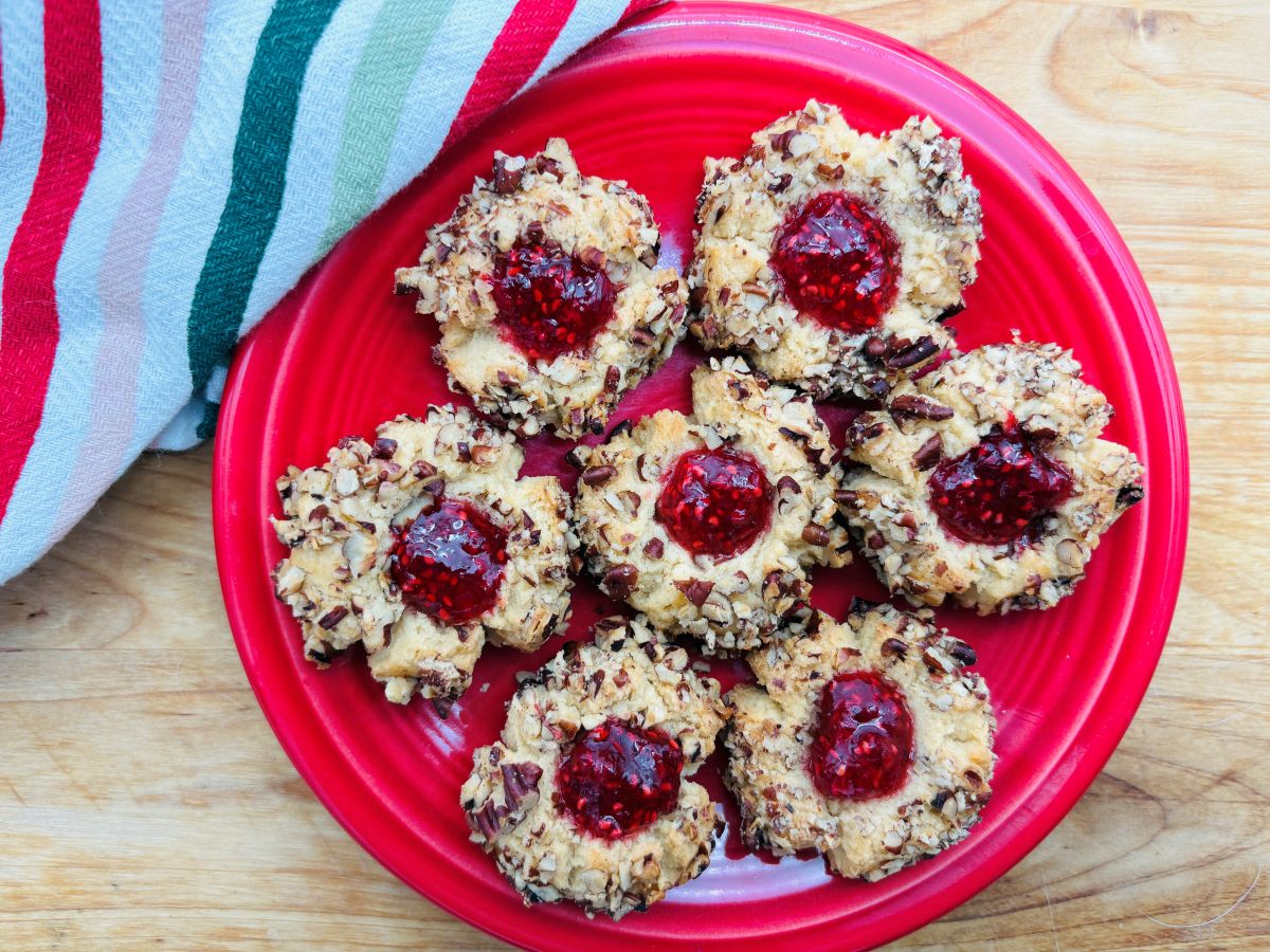 A red plate holds seven thumbprint cookies coated in chopped nuts and filled with red jam, placed on a wooden surface next to a striped towel.