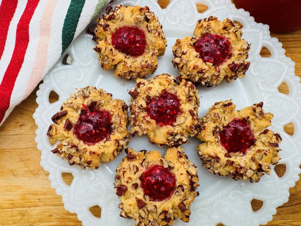 A white plate holds six thumbprint cookies topped with red jam and surrounded by chopped nuts. The plate sits on a wooden surface next to a red, green, and white striped cloth.