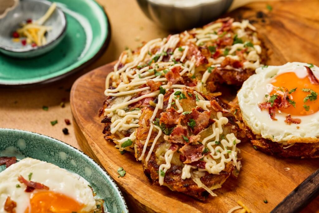 A wooden board topped with crispy potato pancakes drizzled with creamy sauce, sprinkled with bacon and herbs, next to a sunny-side-up egg. Plates with similar food are nearby.