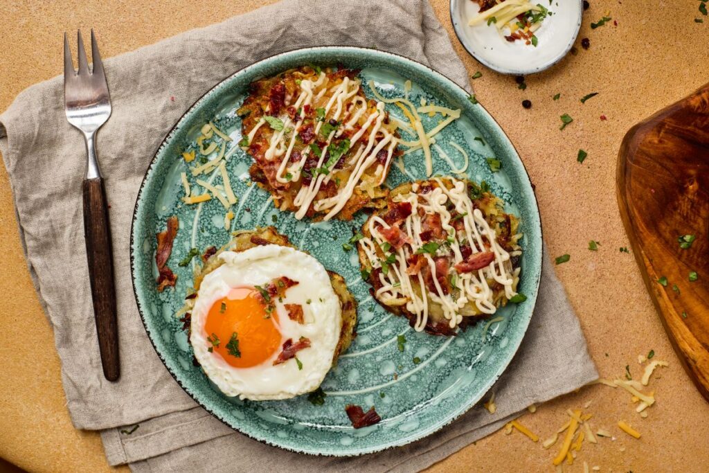 A blue plate with three potato pancakes topped with shredded meat, sauce, and herbs; one pancake has a sunny side up egg. Plate is on a beige napkin with a fork beside it. Small bowl of cheese and herbs in the background.