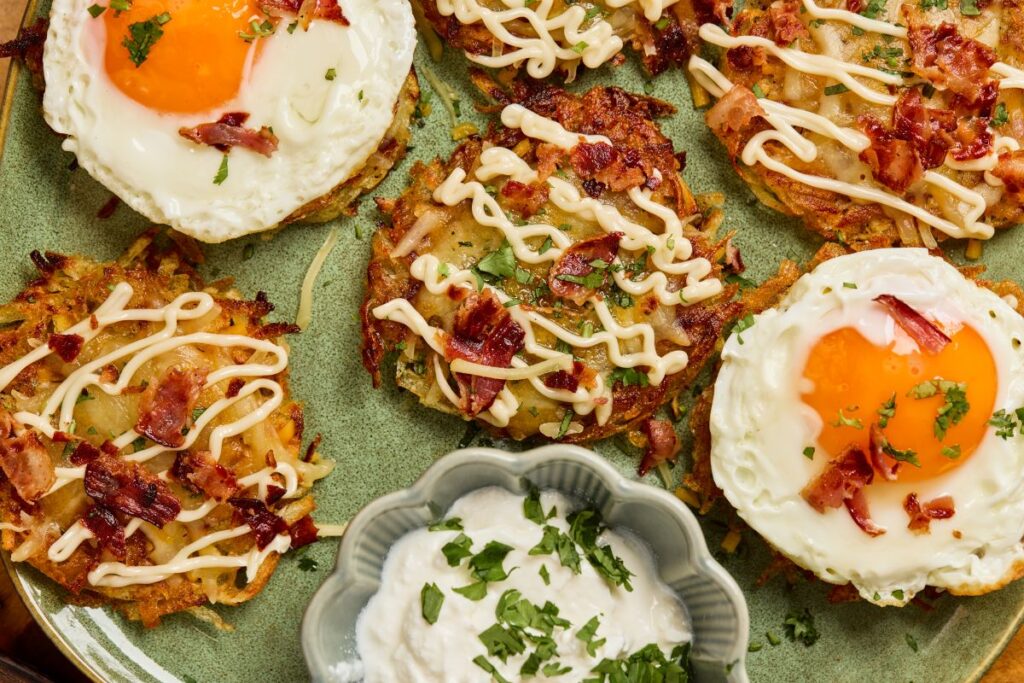 A plate of crispy hash browns topped with fried eggs, chopped bacon, drizzled mayo, and herbs, with a dish of creamy dipping sauce garnished with parsley in the center.