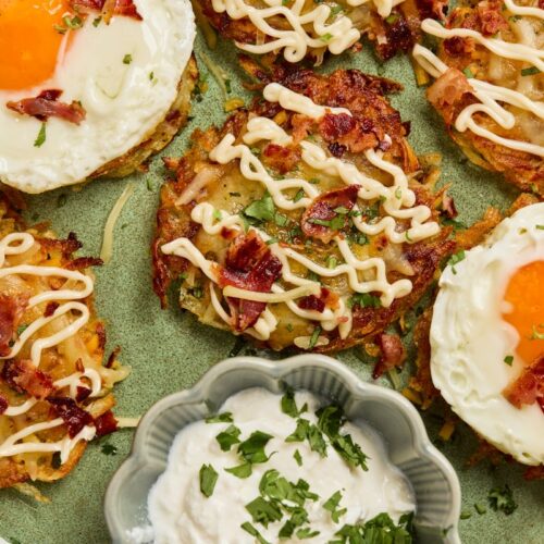 A plate of crispy hash browns topped with fried eggs, chopped bacon, drizzled mayo, and herbs, with a dish of creamy dipping sauce garnished with parsley in the center.
