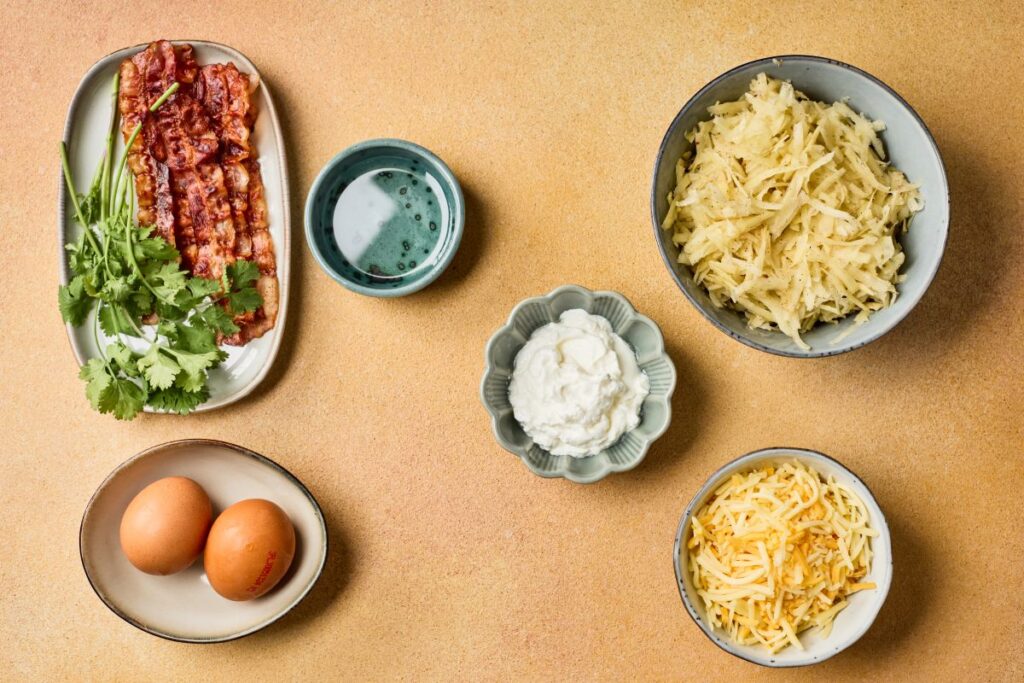 A flat lay of ingredients on a tan surface: a small bowl of grated potatoes, a bowl of shredded cheese, a plate with cilantro and cooked bacon strips, two eggs, a bowl of sour cream, and a small dish of oil.