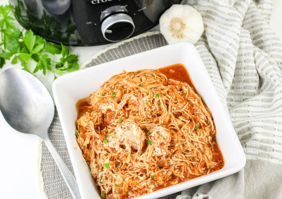 A square white bowl filled with shredded chicken in tomato sauce, garnished with herbs. Nearby are a spoon, a garlic bulb, a striped towel, fresh parsley, and part of a slow cooker.