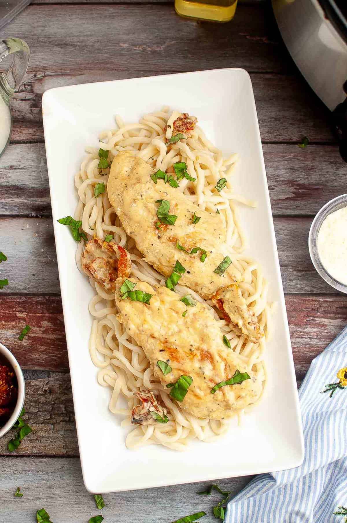 A rectangular white plate holds two creamy chicken breasts on a bed of noodles, garnished with chopped fresh basil. The plate is on a rustic wooden table with parmesan, sun-dried tomatoes, and a slow cooker nearby.