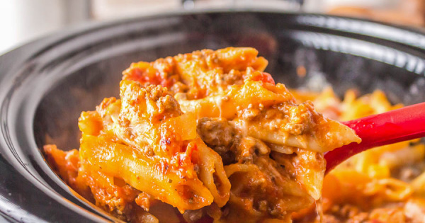 A close-up of cheesy baked ziti with ground meat and tomato sauce being lifted from a slow cooker with a red spoon.