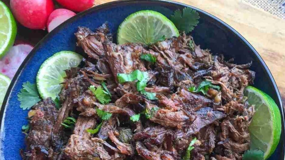 A blue bowl filled with shredded, seasoned beef garnished with fresh cilantro and lime slices, surrounded by whole radishes and lime halves on a wooden surface.