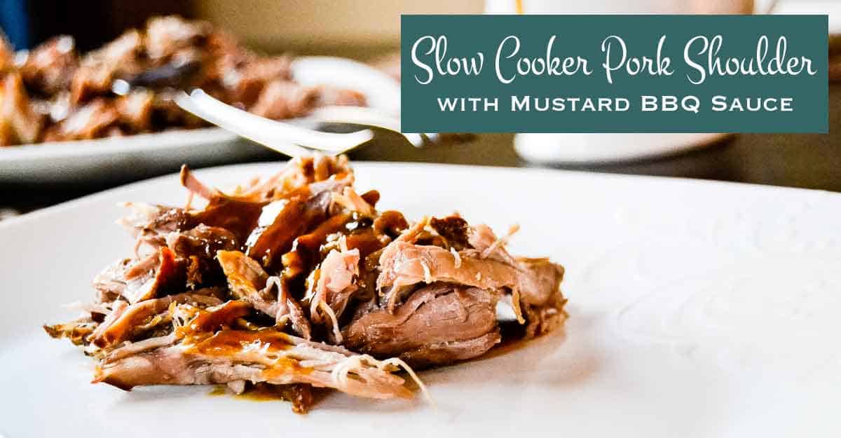 A close-up of shredded slow cooker pork shoulder with mustard BBQ sauce on a white plate. In the background, more pork is visible on another plate. Text overlay reads: “Slow Cooker Pork Shoulder with Mustard BBQ Sauce.”.