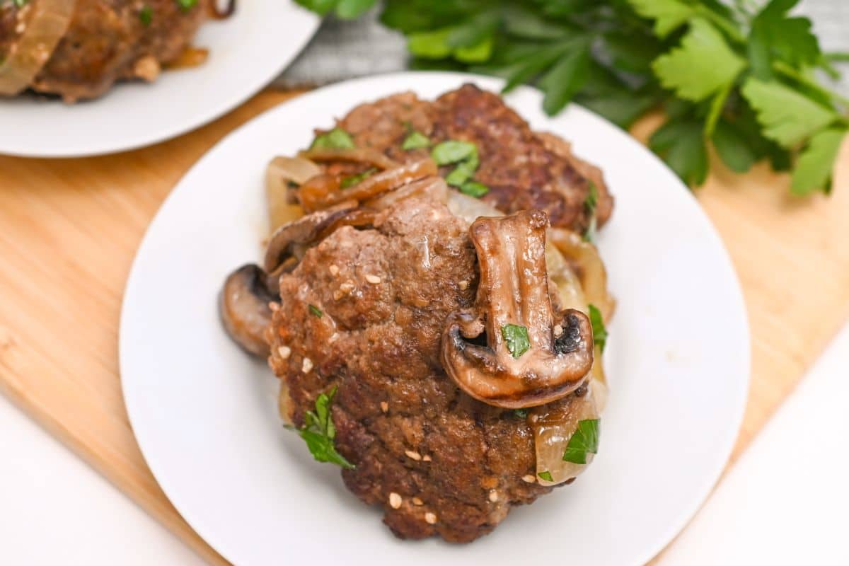 A white plate with cooked Salisbury steak topped with sautéed mushrooms and onions, garnished with chopped parsley. Fresh parsley is visible in the background on a wooden surface.