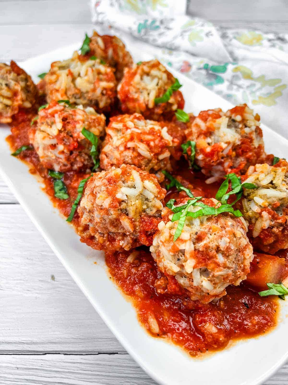 A white rectangular plate with several rice-stuffed meatballs topped with tomato sauce and fresh chopped herbs, arranged on a light wooden surface. A floral napkin is partially visible in the background.