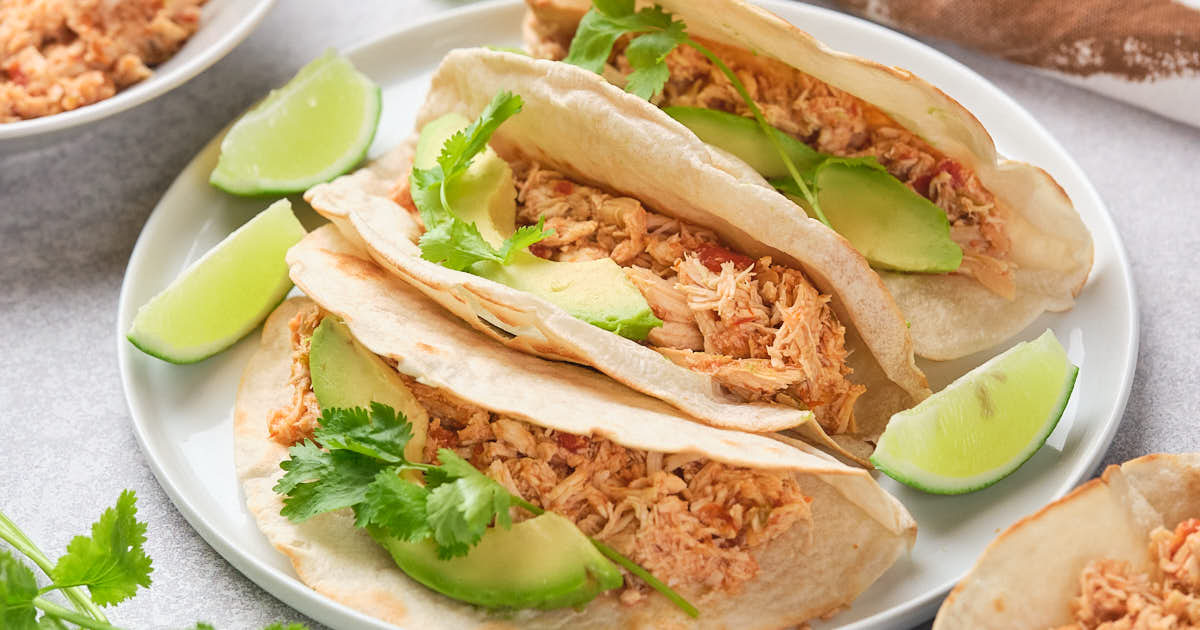 Three soft tacos filled with shredded chicken, avocado slices, and cilantro are arranged on a plate, garnished with lime wedges. A bowl with extra chicken and fresh cilantro are visible in the background.