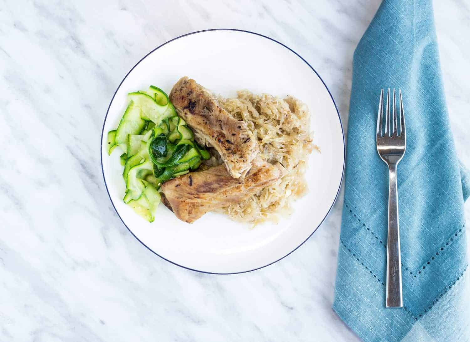 A white plate with two pork ribs, sautéed zucchini ribbons, and a serving of sauerkraut, placed next to a blue cloth napkin and a fork on a marble surface.