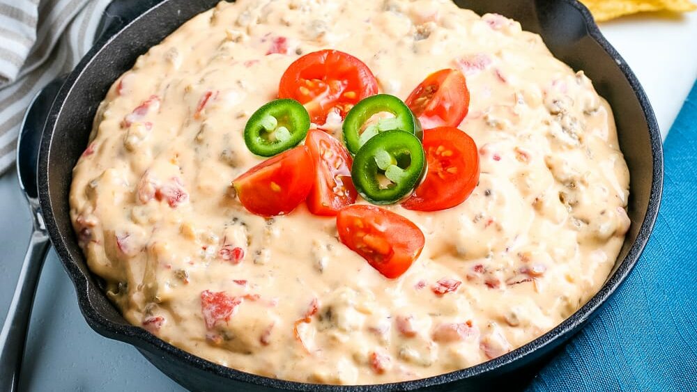 A skillet filled with cheesy dip containing ground meat and tomatoes, topped with sliced cherry tomatoes and jalapeño peppers.