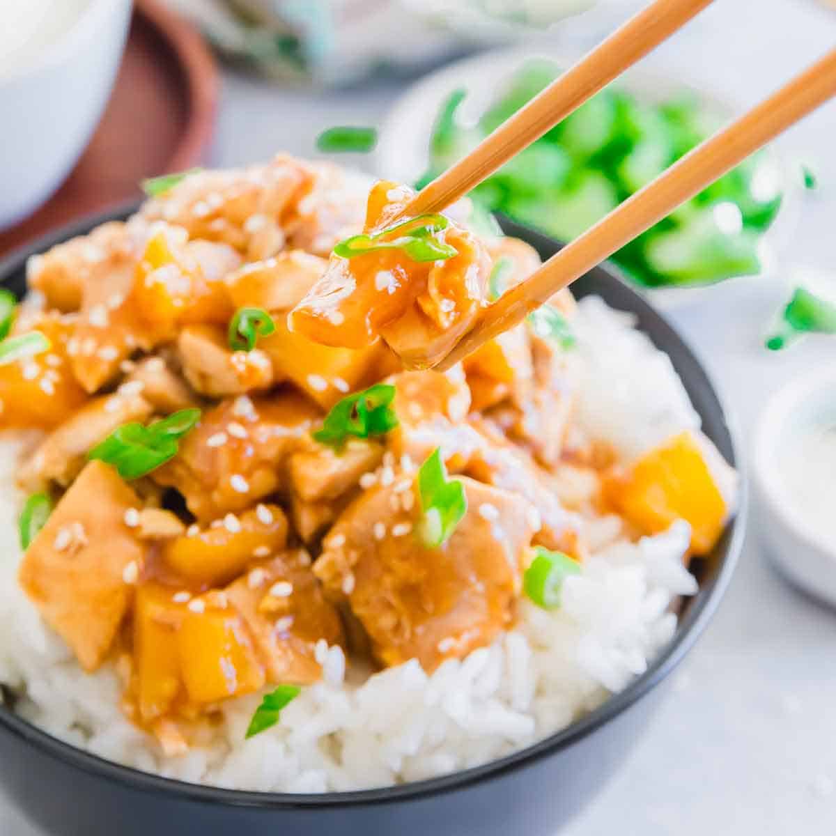 A bowl of white rice topped with orange chicken, diced vegetables, green onions, and sesame seeds. Chopsticks are picking up a piece of chicken, with blurred garnish in the background.