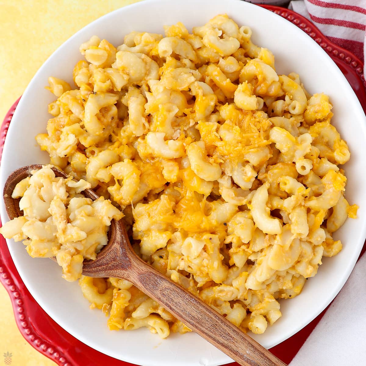A bowl of creamy macaroni and cheese sits on a red plate. A wooden spoon rests in the bowl, scooping up some of the cheesy pasta. The dish appears rich and golden with melted cheese throughout.