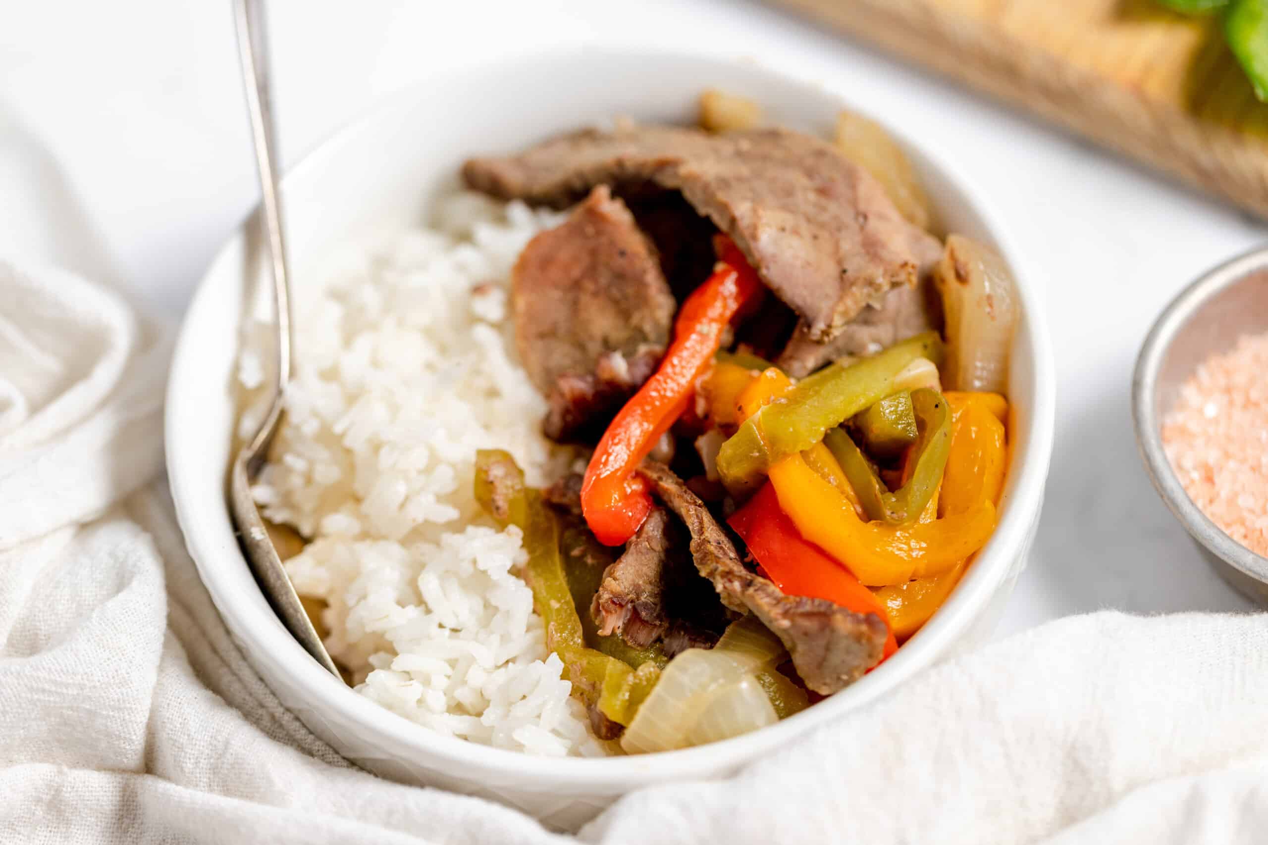 A white bowl filled with white rice, cooked beef strips, and sautéed bell peppers and onions, with a fork resting inside the bowl.