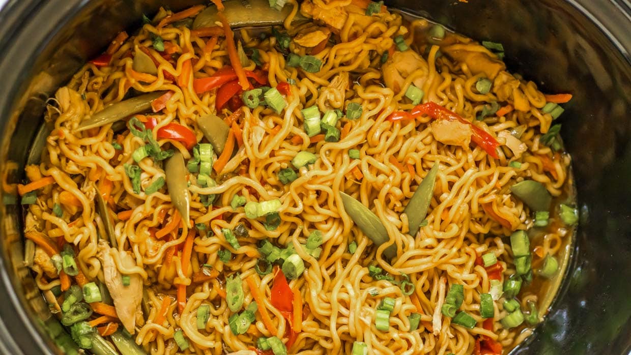 A close-up of cooked noodles mixed with vegetables like chopped green onions, red peppers, and carrots, all in a savory sauce inside a black pot or slow cooker.