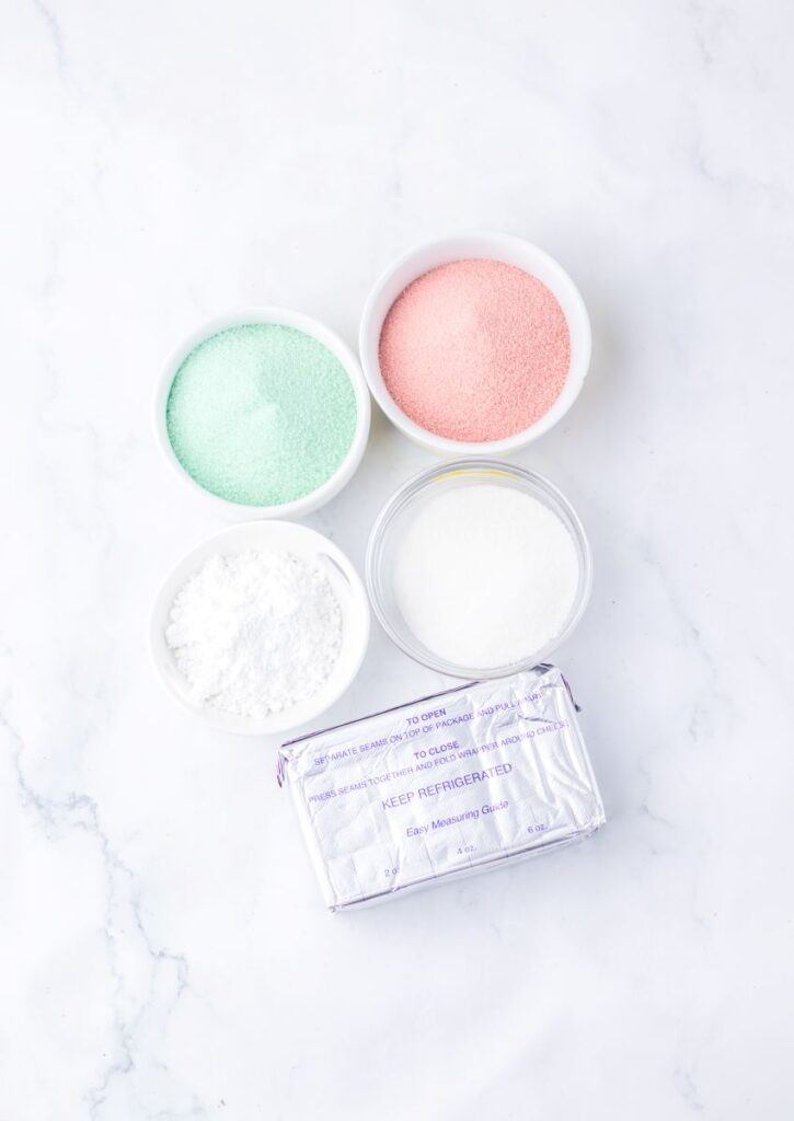 Four small bowls contain pastel green, pastel pink, and white powders, along with a block of cream cheese in silver foil, all arranged on a white marble surface.