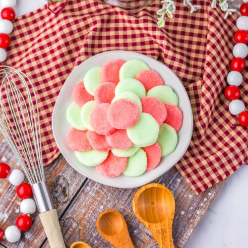 A bowl of pink and green Jello candies on a wooden board, surrounded by wooden spoons, a whisk, red and white bead garland, and a red checkered cloth.