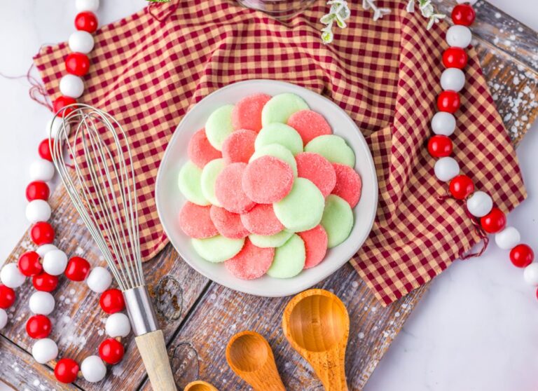 A bowl of pink and green Jello candies on a wooden board, surrounded by wooden spoons, a whisk, red and white bead garland, and a red checkered cloth.