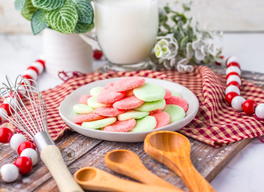 A plate of pink and green jello candies sits on a wooden board with wooden spoons, a whisk, a red plaid cloth, a glass of milk, and festive decorations nearby.