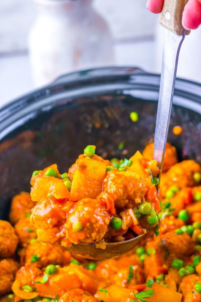 A ladle scoops up a hearty stew with meatballs, potatoes, peas, and carrots in a rich orange sauce from a slow cooker.