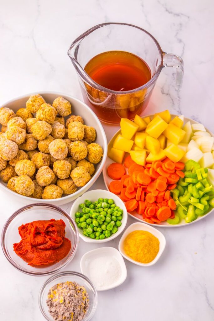 A variety of ingredients on a white surface, including a bowl of small uncooked meatballs, chopped potatoes, carrots, celery, onion, peas, tomato paste, broth in a measuring cup, seasoning powders, and salt.