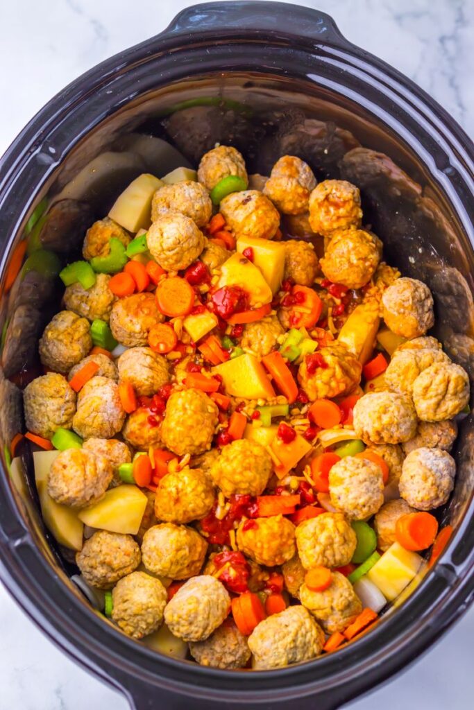 A slow cooker filled with raw meatballs, chopped carrots, potatoes, celery, and diced red peppers, ready to be cooked. The colorful ingredients are layered evenly inside the pot.