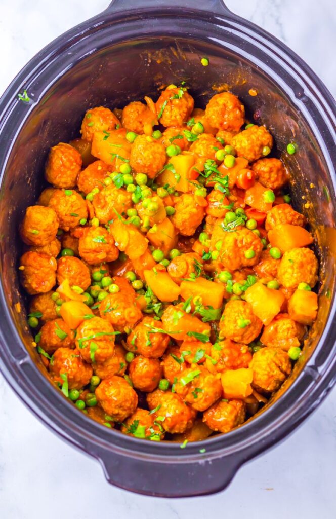 A slow cooker filled with saucy meatballs, yellow bell peppers, green peas, and chopped herbs, all mixed together in a vibrant orange sauce.